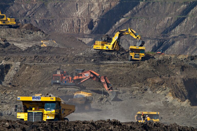 A wide-angle, high-perspective photograph of a vast open-pit mining operation. Multiple pieces of heavy industrial machinery are active across tiered dirt terraces, including large yellow haul trucks and hydraulic excavators in yellow and orange. The scene captures the scale of resource extraction, with steep, excavated rock walls in the background and a dusty, industrial atmosphere.