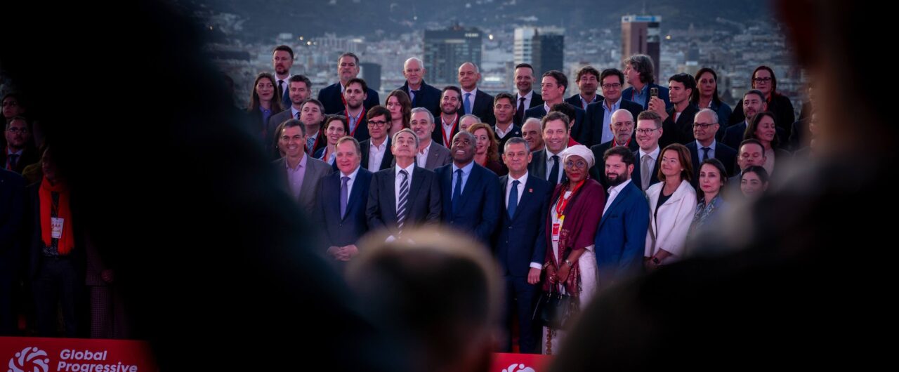 A large group photo of numerous participants from the Global Progressive Mobilisation conference, posing in formal attire against the evening skyline of Barcelona. While the individuals in the center look toward the camera, blurred silhouettes in the foreground frame the shot. The background features the dense urban landscape and the prominent Torre de Collserola on the hills under a twilight sky; a red event banner with the organization's logo is visible at the bottom of the frame.