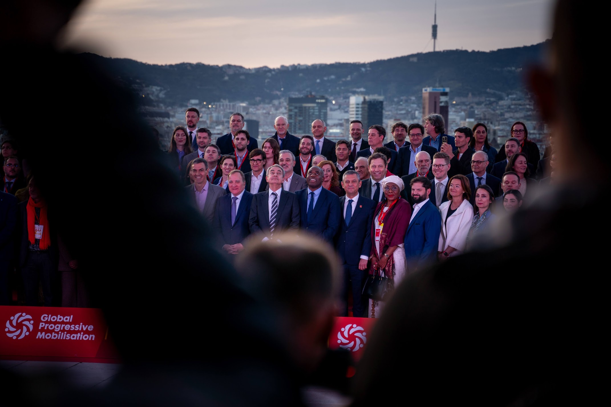 A large group photo of numerous participants from the Global Progressive Mobilisation conference, posing in formal attire against the evening skyline of Barcelona. While the individuals in the center look toward the camera, blurred silhouettes in the foreground frame the shot. The background features the dense urban landscape and the prominent Torre de Collserola on the hills under a twilight sky; a red event banner with the organization's logo is visible at the bottom of the frame.