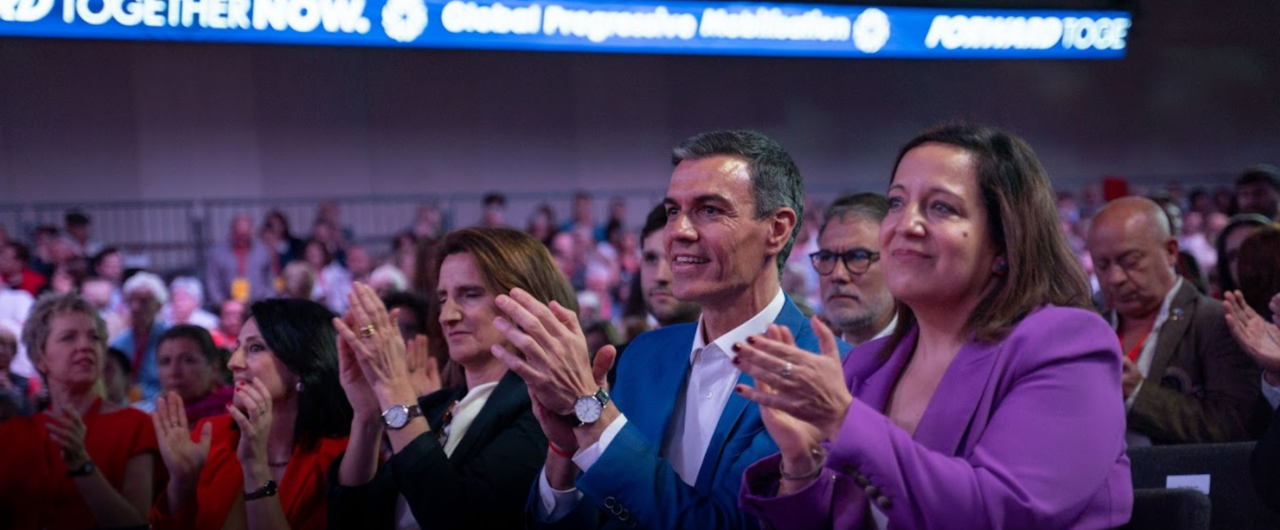 Spanish Prime Minister Pedro Sánchez sits in the front row of the Global Progressive Mobilisation Summit, smiling and applauding alongside other attendees. Sánchez, dressed in a bright blue suit, is flanked by several colleagues, including a woman in a purple blazer and another in a dark jacket. Above them, a long digital banner illuminates the venue with the slogans "Global Progressive Mobilisation" and "FORWARD TOGETHER NOW." The scene captures an energetic moment of unity and support within a crowded conference hall.