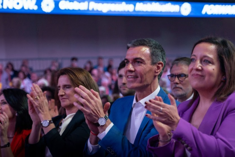 Spanish Prime Minister Pedro Sánchez sits in the front row of the Global Progressive Mobilisation Summit, smiling and applauding alongside other attendees. Sánchez, dressed in a bright blue suit, is flanked by several colleagues, including a woman in a purple blazer and another in a dark jacket. Above them, a long digital banner illuminates the venue with the slogans "Global Progressive Mobilisation" and "FORWARD TOGETHER NOW." The scene captures an energetic moment of unity and support within a crowded conference hall.