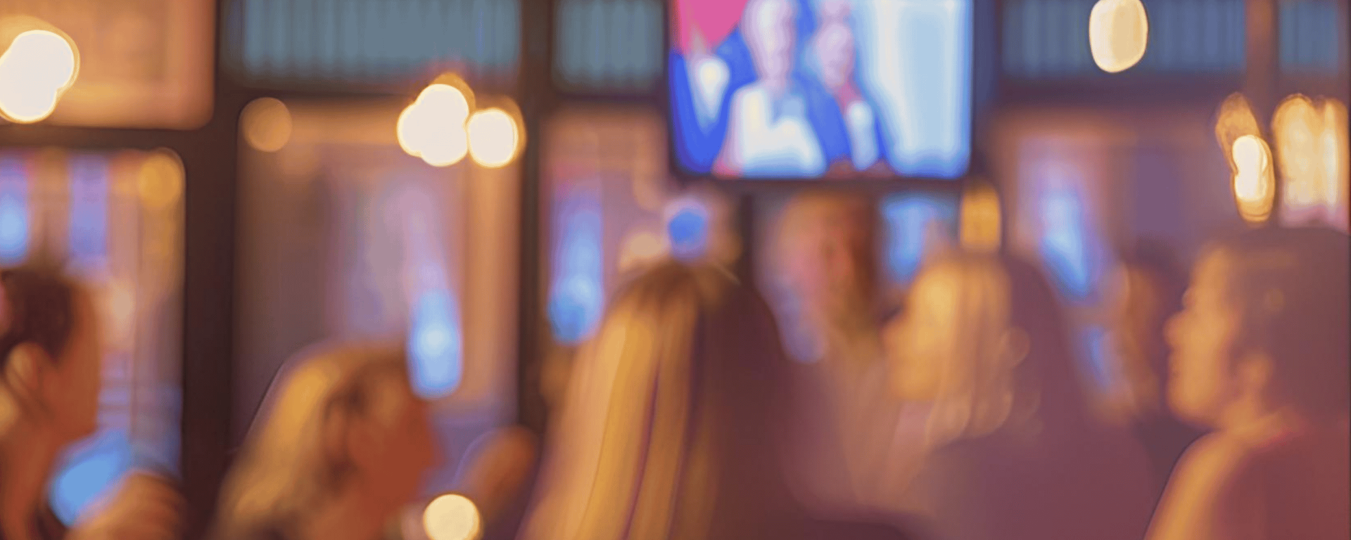 Verschwommene Aufnahme einer Menschenmenge in einer Bar oder einem Veranstaltungsraum mit warmem Licht. Im Hintergrund läuft eine Fernsehsendung auf einem Bildschirm, die eine politische Debatte oder eine Nachrichtensendung zeigen könnte. Die Personen scheinen in Gespräche vertieft oder auf den Bildschirm fokussiert zu sein.