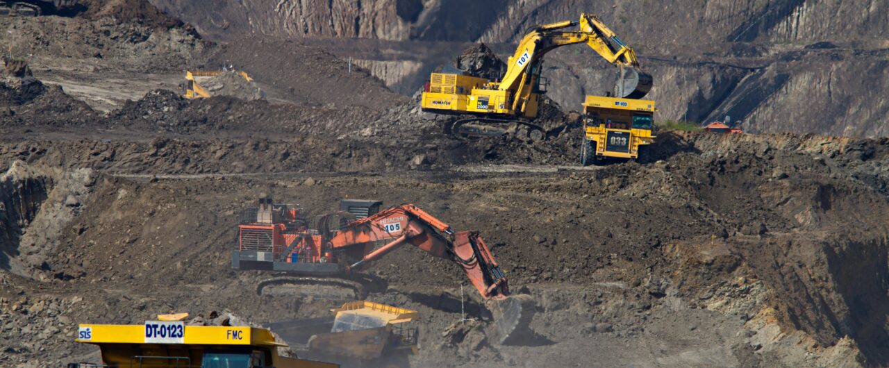A wide-angle, high-perspective photograph of a vast open-pit mining operation. Multiple pieces of heavy industrial machinery are active across tiered dirt terraces, including large yellow haul trucks and hydraulic excavators in yellow and orange. The scene captures the scale of resource extraction, with steep, excavated rock walls in the background and a dusty, industrial atmosphere.