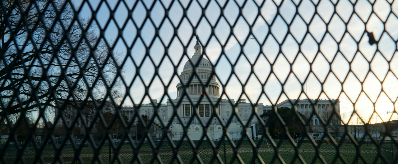 Das US-Kapitol in Washington D.C. steht im Zentrum des Bildes unter einem leicht bewölkten Himmel. Die weiße Kuppel des amerikanischen Regierungsgebäudes wird im Vordergrund durch die rautenförmigen Maschen eines dunklen Sicherheitsgitters oder Bauzauns hindurch fotografiert, was eine symbolische Barriere erzeugt. Links im Hintergrund sind kahle Bäume zu sehen, während das warme Licht der tiefstehenden Sonne die Szene seitlich beleuchtet.