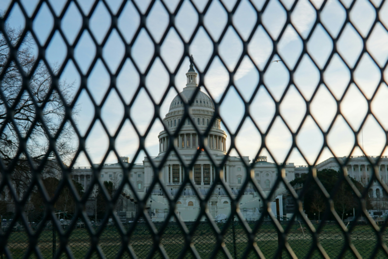 Das US-Kapitol in Washington D.C. steht im Zentrum des Bildes unter einem leicht bewölkten Himmel. Die weiße Kuppel des amerikanischen Regierungsgebäudes wird im Vordergrund durch die rautenförmigen Maschen eines dunklen Sicherheitsgitters oder Bauzauns hindurch fotografiert, was eine symbolische Barriere erzeugt. Links im Hintergrund sind kahle Bäume zu sehen, während das warme Licht der tiefstehenden Sonne die Szene seitlich beleuchtet.