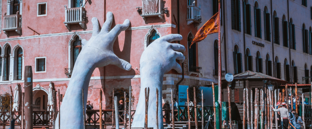 Großformatige weiße Skulptur zweier riesiger Hände von Lorenzo Quinn, die aus dem Canal Grande in Venedig ragen und die Fassade des historischen Hotels Ca' Sagredo zu stützen scheinen, als Symbol für den Klimawandel und die Kunstfreiheit.
