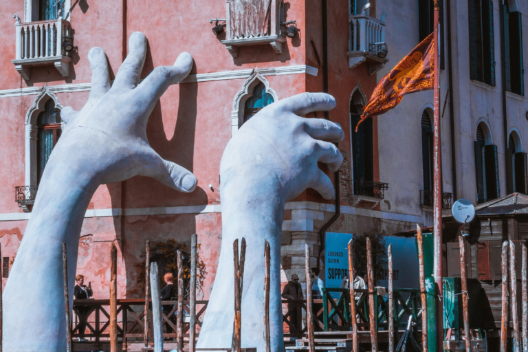 Großformatige weiße Skulptur zweier riesiger Hände von Lorenzo Quinn, die aus dem Canal Grande in Venedig ragen und die Fassade des historischen Hotels Ca' Sagredo zu stützen scheinen, als Symbol für den Klimawandel und die Kunstfreiheit.