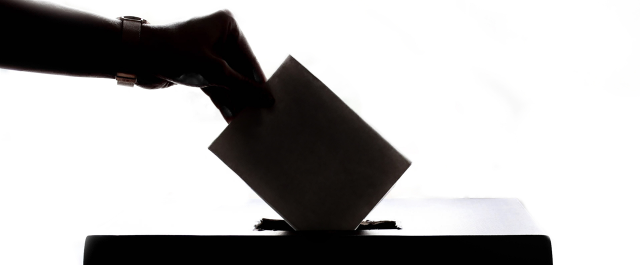 Silhouette of a hand wearing a wristwatch inserting a folded ballot paper into the slot of a ballot box against a bright white background, symbolizing voting, democracy, and elections.