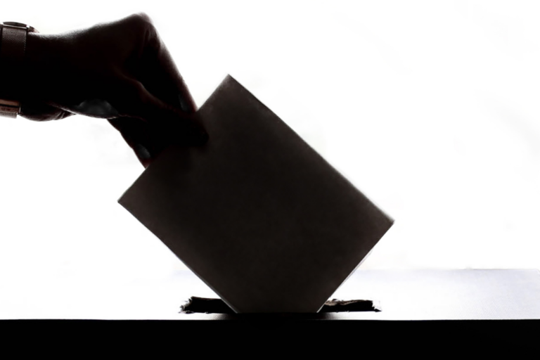 Silhouette of a hand wearing a wristwatch inserting a folded ballot paper into the slot of a ballot box against a bright white background, symbolizing voting, democracy, and elections.