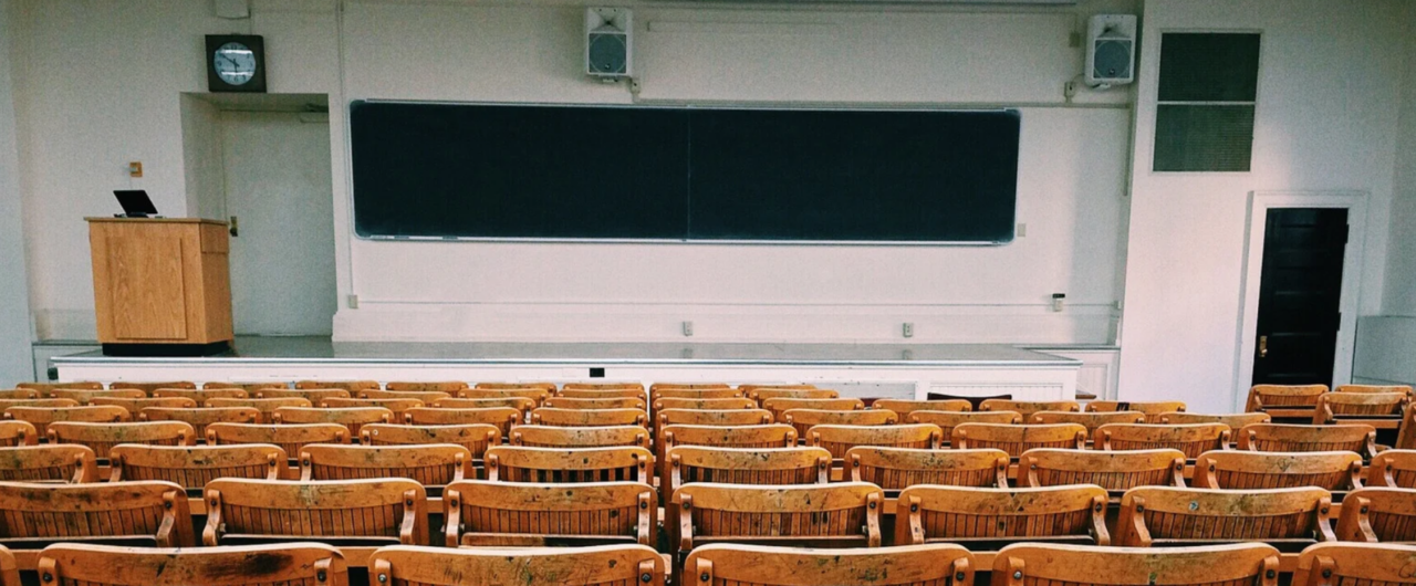 Ein leerer Hörsaal einer Universität mit Reihen aus hölzernen Klappstühlen im Vordergrund, einem Dozentenpult auf einer kleinen Bühne und einer großen dunklen Wandtafel an der Frontseite, ideal als Symbol für Hochschulbildung, Vorlesungen und wissenschaftliche Lehre.