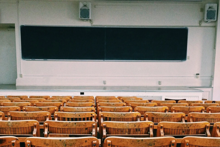 Ein leerer Hörsaal einer Universität mit Reihen aus hölzernen Klappstühlen im Vordergrund, einem Dozentenpult auf einer kleinen Bühne und einer großen dunklen Wandtafel an der Frontseite, ideal als Symbol für Hochschulbildung, Vorlesungen und wissenschaftliche Lehre.