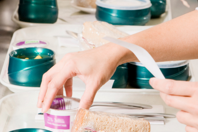 Mitarbeitende im Bereich Gemeinschaftsverpflegung und Care Catering bereiten in einer professionellen Küchenumgebung Mahlzeiten vor und portionieren Speisen auf Tabletts. Zu sehen sind verpackte Lebensmittel, Besteck und Mehrwegbehälter, die für die Auslieferung oder Versorgung in Krankenhäusern, Pflegeeinrichtungen oder sozialen Einrichtungen vorbereitet werden.